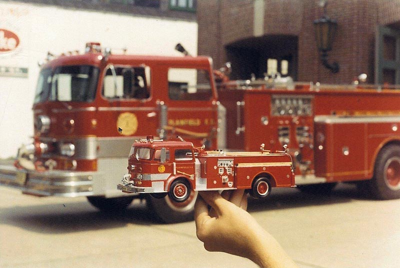 Tom holds up one of his commissioned scale model fire trucks in front of the full-size engine that it was patterned after. 